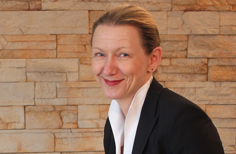 Portrait of Dagmar Saschek in front of a beige sandstone wall.
